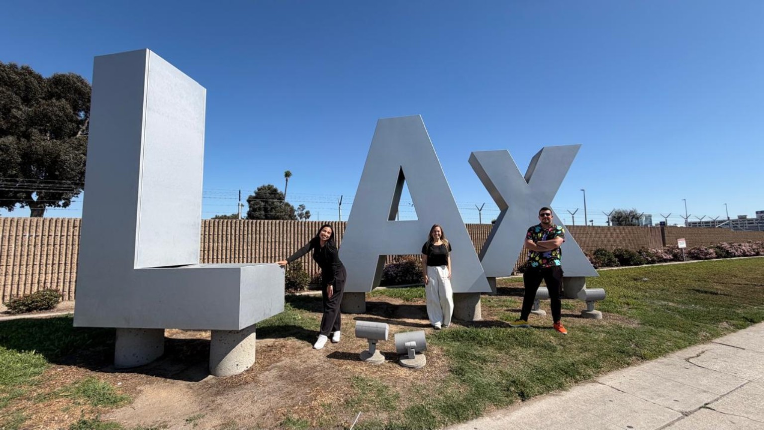 Três pessoas diante do letreiro LAX, em Los Angeles